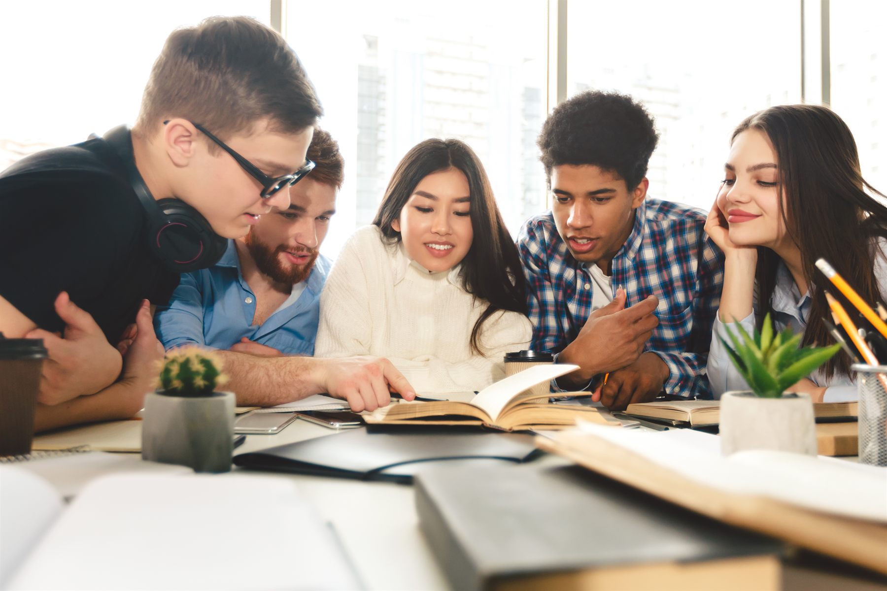 A diverse group of students studying together around a table, reading and discussing an open book.