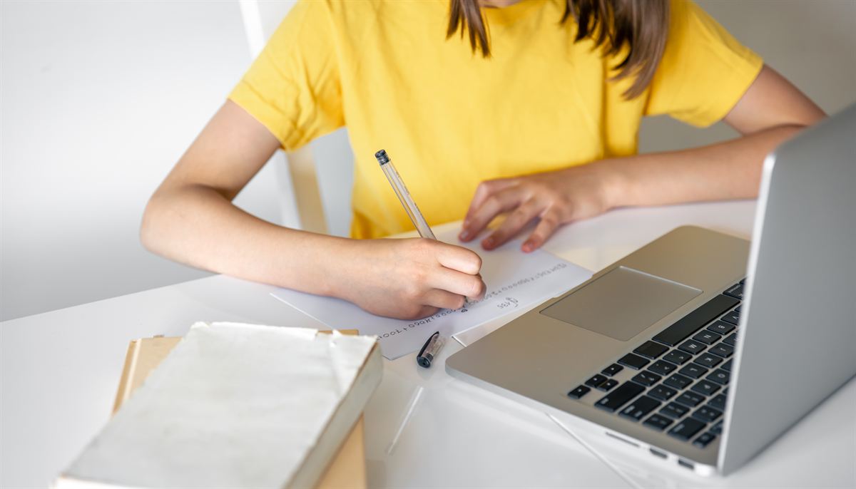 A student writing notes on paper beside an open laptop.